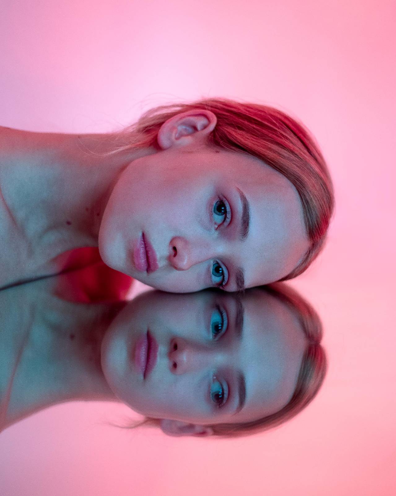 Close-up of a girl's face leaning on a mirror, creating a reflection, with a pink backdrop, photographed in Kolding.